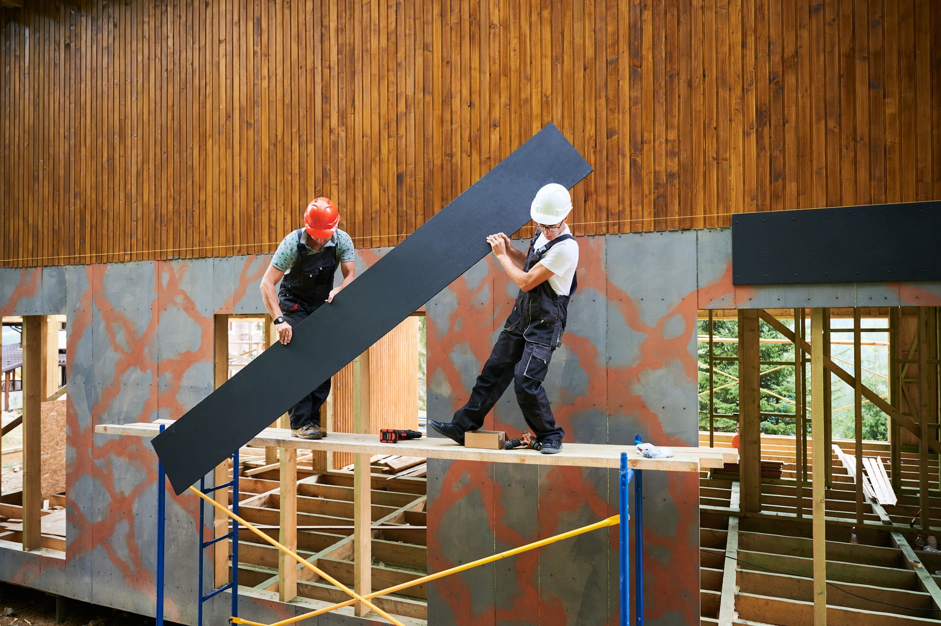 Workers cladding facade of house with cement particle boards while constructing wooden frame house.